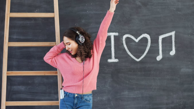 Chica escuchando música con auriculares, levantando la mano mientras baila, con una decoración de “I love music” al fondo